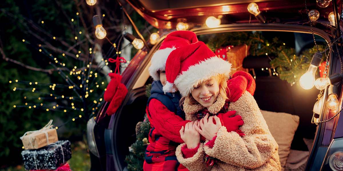A cheerful holiday scene with two people in Santa hats hugging by a car trunk, adorned with lights and gifts, evoking warmth and festive joy.