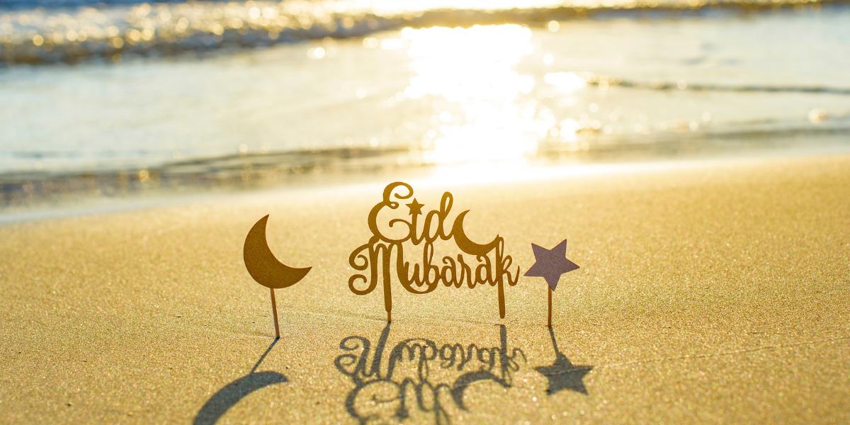 "Eid Mubarak sign with crescent and star on golden sand, reflecting sunlight. Gentle waves shimmer in the background, conveying warmth and celebration."