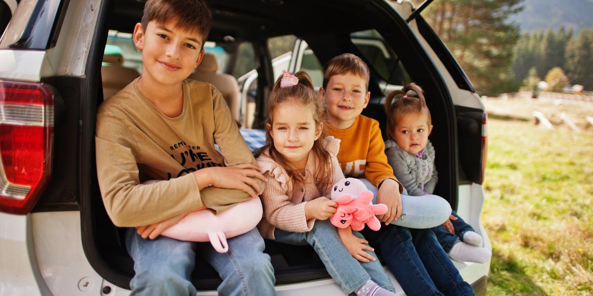Four young children sit happily in the open trunk of a car, surrounded by nature. They smile warmly, each holding a plush toy, conveying joy and togetherness.