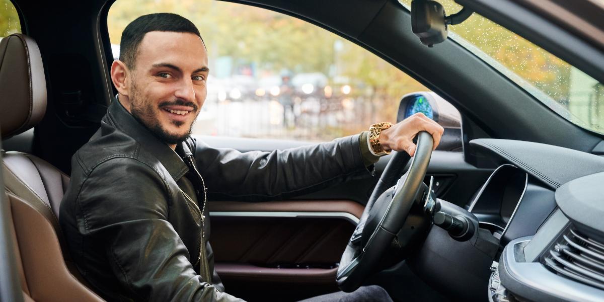 A smiling man sits in the driver's seat of a car, wearing a black leather jacket. The outside view shows blurred trees, suggesting motion.