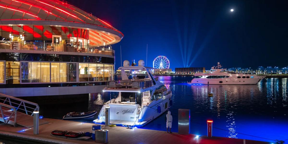 A vibrant harbor at night, featuring a luxurious yacht docked beside a modern, illuminated building. A Ferris wheel and city lights illuminate the background.